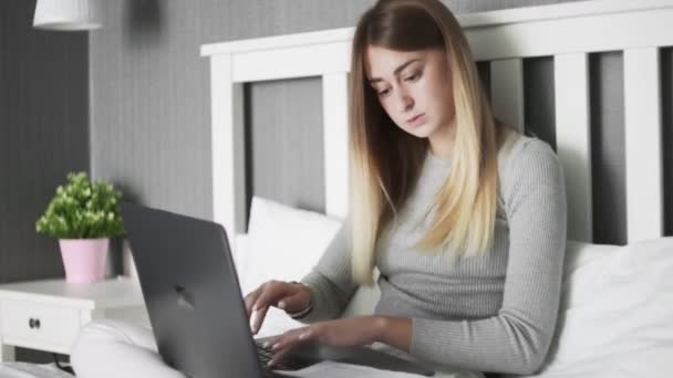 Серйозна жінка сидить на ліжку, використовує ноутбук і працює з документами — стокове відео