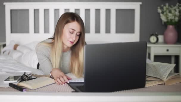 Una joven estudiante escribe en un cuaderno. En la cama de aprendizaje a distancia para los estudiantes. Coronavirus. Cuarentena. Una mujer mira una computadora portátil. freelancer, trabajo a distancia, trabajo en casa, educación en línea — Vídeos de Stock