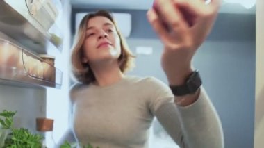 Buzdolabının İçinden Dışarıya Bakış Kadın kapıyı açarken bir elmayı alır ve geri koyar ve çöreği alır.