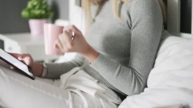 Yatağa uzanmış, kahve içen ve tablet kullanan bir kadın.