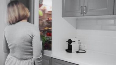 Başı ağrıyan ya da akşamdan kalma bir kadın mutfaktaki buzdolabının kapısını açar ve bir şişe su alır.