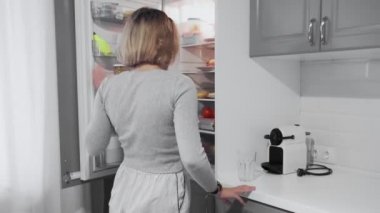 Kadın mutfaktaki buzdolabının kapısını açıyor ve bir şişe süt alıyor, sonra da bardağa süt döküyor.