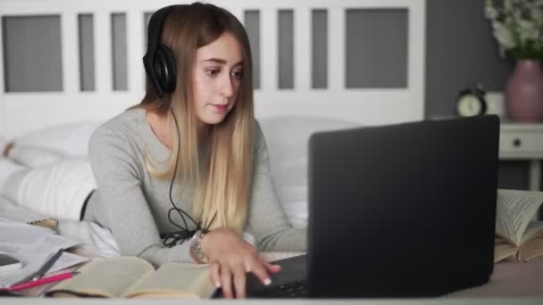 Jovem estudante com fones de ouvido deitado na cama usando notebook. Ensino à distância para estudantes . — Vídeo de Stock