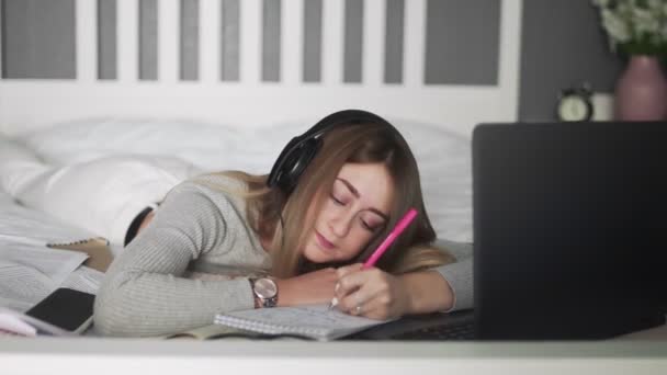 Jonge vrouw met koptelefoon liggend op het bed en het maken van hotes op notebook — Stockvideo