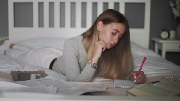 Молода студентка лежить на ліжку і робить записку в блокноті — стокове відео