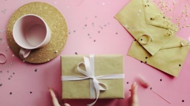 Kadın eli, beyaz fiyonklu el işi kağıdına sarılı Noel hediyesini alıp geri koyuyor. Tahta, pembe masa, düz uzanma.