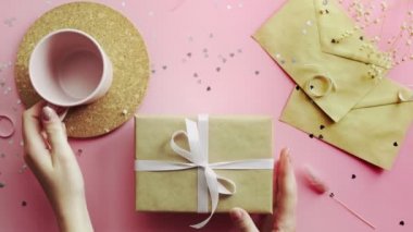 Kadın elleri boş bir bardak ve beyaz kurdeleyle sarılmış bir Noel hediyesi alıyor. Tahta, pembe masa, düz uzanma.