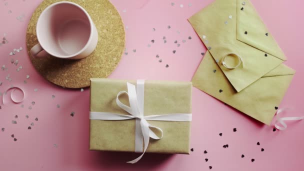Manos de mujer tomando un regalo de Navidad envuelto en papel artesanal con lazo blanco. Vista superior sobre tabla rosada de madera, posición plana — Vídeos de Stock