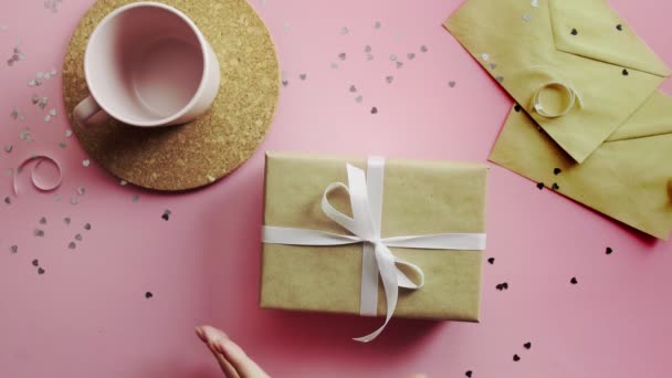 白い弓でクラフト紙に包まれたクリスマスの贈り物を振る女性の手。木製のピンクのテーブルの上からの眺め、フラットレイアウト — ストック動画