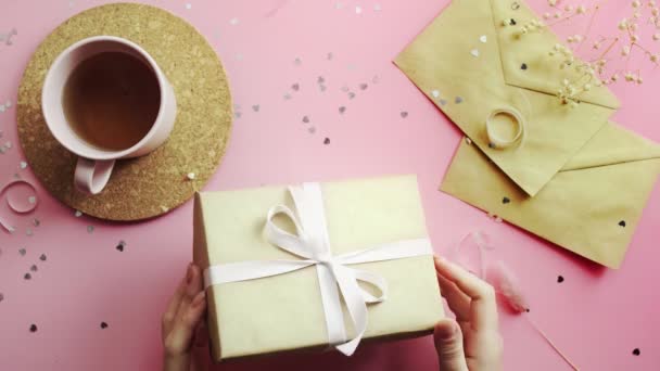 Les mains féminines attachent un cadeau de Noël enveloppé dans du papier artisanal avec un arc blanc. Vue du dessus sur table rose bois, plan plat — Video