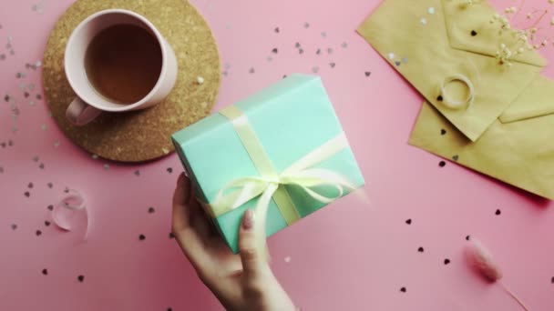 Des mains de femme serrant un cadeau de Noël enveloppé dans un papier vert avec un arc jaune. Vue du dessus sur table rose bois, plan plat — Video