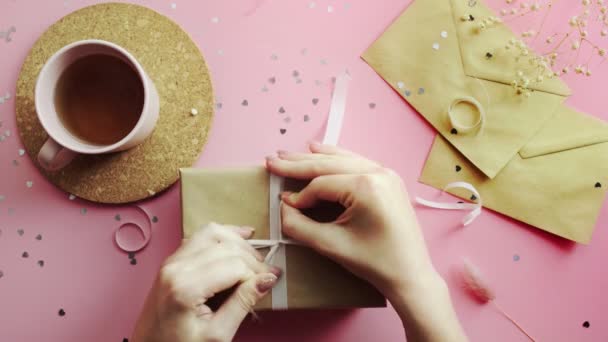 Vrouwelijke handen strikken strik een kerstcadeau verpakt in ambachtelijk papier met witte strik. Bovenaanzicht op hout roze tafel, plat gelegd — Stockvideo