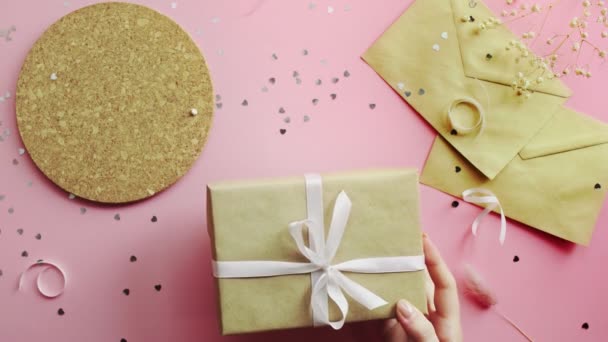 Les mains féminines attachent un cadeau de Noël enveloppé dans du papier artisanal avec un arc blanc. Vue du dessus sur table rose bois, plan plat — Video