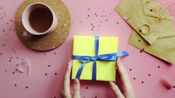 Manos de mujer agitando un regalo de Navidad envuelto en un papel amarillo con lazo azul. Vista superior sobre tabla rosada de madera, posición plana — Vídeo de stock