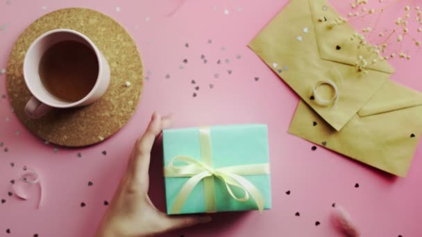 Mano de mujer sacudiendo un regalo de Navidad envuelto en un papel verde con lazo amarillo. Vista superior sobre tabla rosada de madera, posición plana — Vídeos de Stock