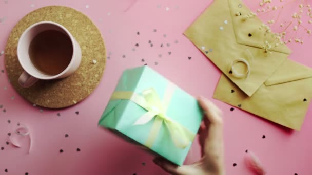 Женские руки осматривают и трясут рождественский подарок, завернутый в зеленую бумагу с желтым бантом. Вид сверху на деревянный розовый стол, плоский — стоковое видео