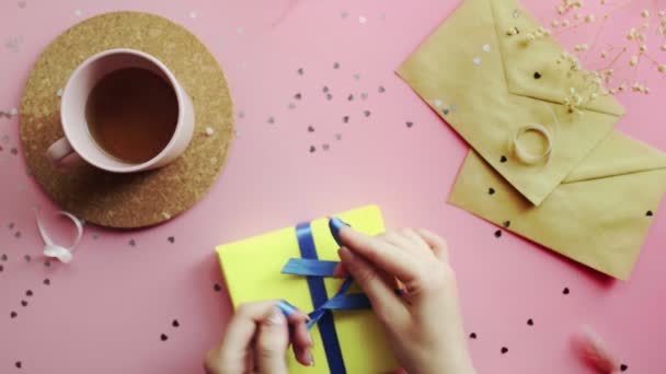 Mains féminines attachant un arc bleu sur un cadeau de Noël enveloppé dans du papier jaune. Vue du dessus sur table rose bois, plan plat — Video