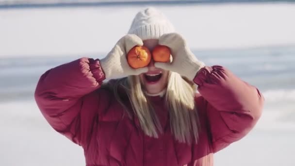 Mujer joven en sombrero de punto blanco y chaqueta de invierno roja posando con dos mandarinas en la cámara — Vídeos de Stock