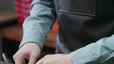 Önlüklü adam, deri atölyesinde kahverengi deri kemer yapmak için iğne kullanıyor. Deri atölyedeki deri kemerin çalışma süreci..