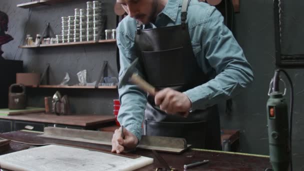 Curtidor profesional haciendo agujeros en un nuevo cinturón de cuero marrón en el taller de cuero. Proceso de trabajo del cinturón de cuero en el taller de cuero . — Vídeo de stock
