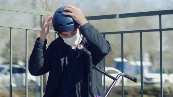 Mladý bmx jezdec v lékařské masce sedící na rampě a v helmě — Stock video