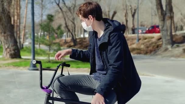 En ung bmx ryttare i medicinsk mask sitter på en cykel och bär hjälm — Stockvideo