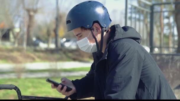 Ένας νεαρός αναβάτης bmx με ιατρική μάσκα κάθεται σε ένα ποδήλατο και χρησιμοποιώντας το smartphone — Αρχείο Βίντεο