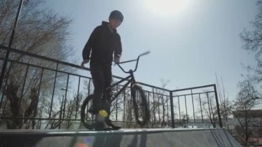 Genç bir BMX sürücüsü rampanın tepesinde duruyor ve ekstrem parktaki ön tekerleği döndürüyor.