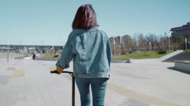Kot ceketli genç bir kadının yaz parkında scooter sürüşünün arka görüntüsü.