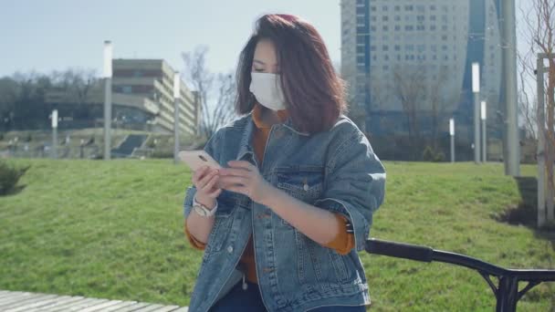 Joven mujer asiática en máscara médica se sienta en el banco y utiliza el teléfono inteligente para grabar un mensaje de voz en el parque de verano — Vídeos de Stock