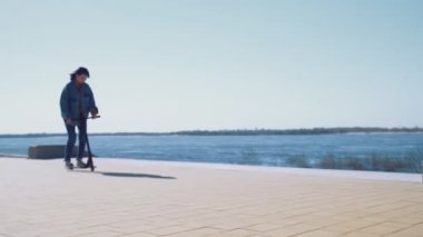 Kot ceketli genç Asyalı kadın nehir kıyısında scooter kullanıyor.