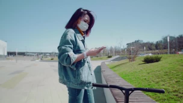 Joven mujer asiática en máscara médica pone el teléfono inteligente en el bolsillo y empezar a paseos en scooter parque de verano — Vídeos de Stock