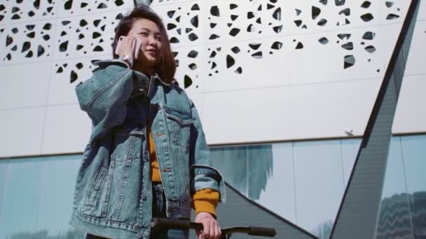 Молодая азиатка в джинсовой куртке гуляет со скутером и разговаривает по телефону в летнем парке — стоковое видео