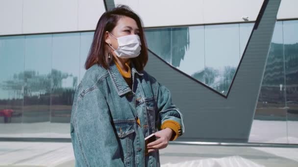 청바지를 입고 의료용 마스크를 쓴 젊은 동양 여성 이 스쿠터를 들고 서서 여름 공원에서 목소리 메시지를 녹음 한다 — 비디오