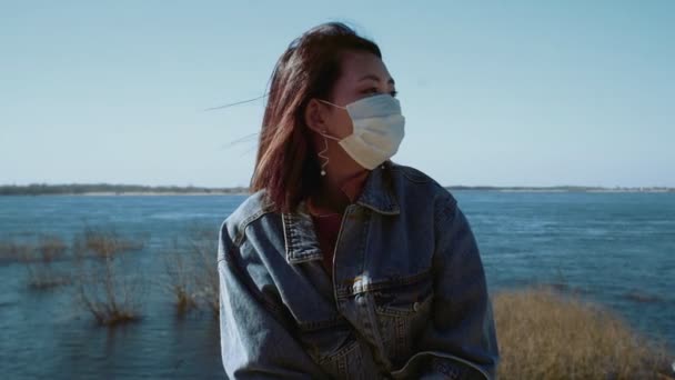 Ung asiatisk kvinna i jeans jacka och medicinsk mask sitter på flodstranden — Stockvideo