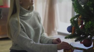 Kazaklı ve kot pantolonlu genç bir kadın yerde oturuyor ve Noel ağacını evde toplarla süslüyor..