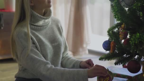 Молодая женщина в свитере и синих джинсах сидит на полу и украшает рождественскую елку мячами дома . — стоковое видео