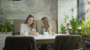 İki genç kadın telefona bakıyor ve kafede gülümsüyor.