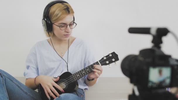 Joven blogger chica atractiva con auriculares en la cabeza está grabando video con cámara, cantando y tocando en la guitarra — Vídeo de stock