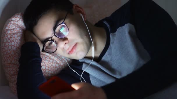 Jeune bel homme hippie utilisant un téléphone intelligent mobile allongé sur le lit à la maison tard dans la nuit. — Video