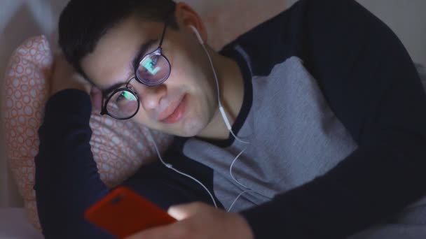 Joven hombre atractivo Hipster utilizando un teléfono inteligente móvil acostado en la cama en casa tarde en la noche . — Vídeo de stock