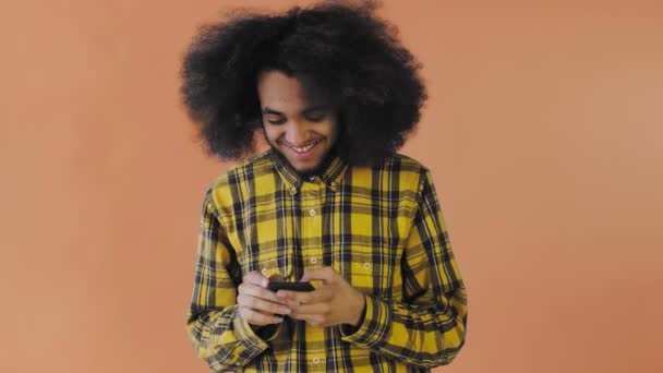 Ritratto di uomo afroamericano con capelli ricci che chatta su smartphone su sfondo arancione. Concetto di emozioni — Video Stock