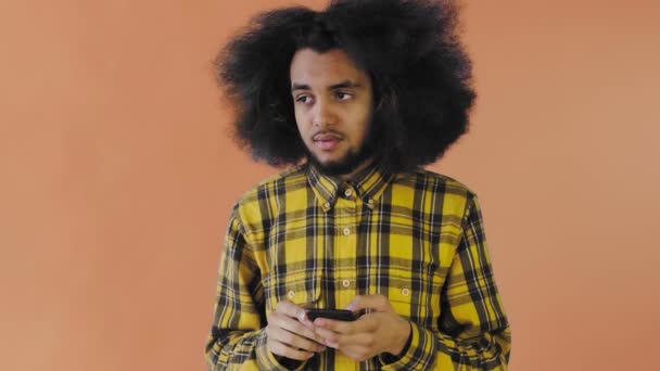 Uomo afroamericano con i capelli ricci sta pensando prima di inviare un messaggio su sfondo arancione. Concetto di emozioni — Video Stock