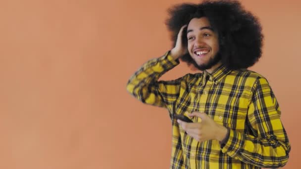 Счастливый привлекательный молодой афроамериканец, пользующийся телефоном и получающий хорошие новости на оранжевом фоне. Концепция эмоций — стоковое видео