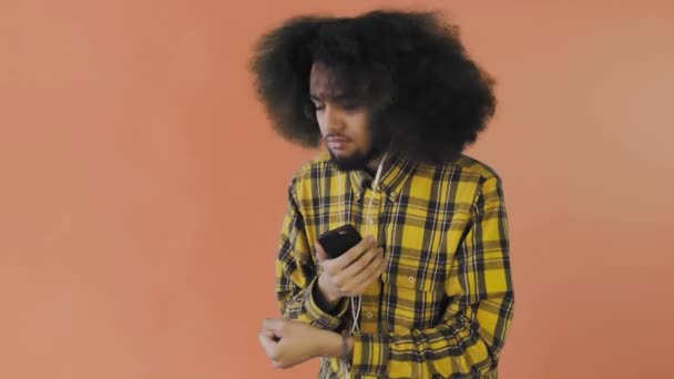Uomo afroamericano positivo che usa il telefono per ascoltare musica nelle cuffie su sfondo arancione. Concetto di emozioni — Video Stock