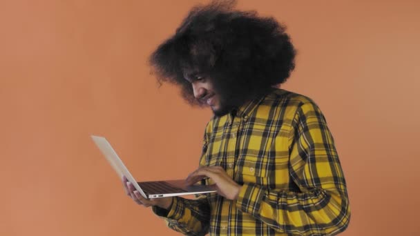 Hombre afroamericano escribiendo en el portátil y sonriendo sobre fondo naranja. Concepto de emociones — Vídeos de Stock