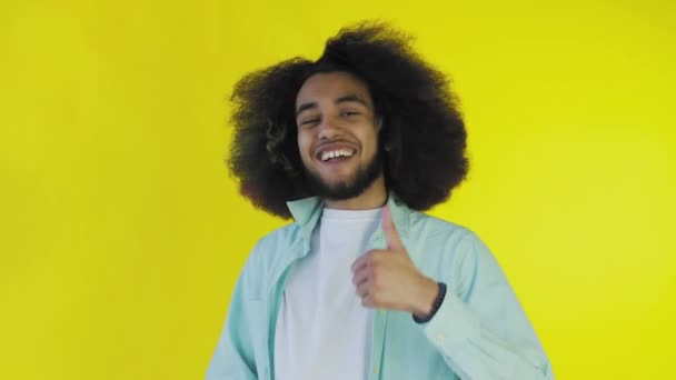 Молодой счастливый афро-американец улыбается, показывая большие пальцы на желтом фоне. Концепция эмоций — стоковое видео
