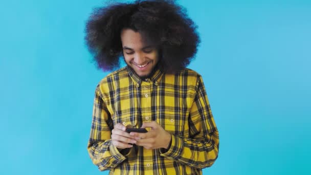 Ritratto di uomo afroamericano con capelli ricci che chatta su smartphone su sfondo blu. Concetto di emozioni — Video Stock