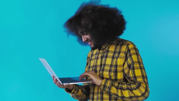 Афроамериканець друкує на ноутбуці і посміхається на синьому фоні. Концепція емоцій — стокове відео
