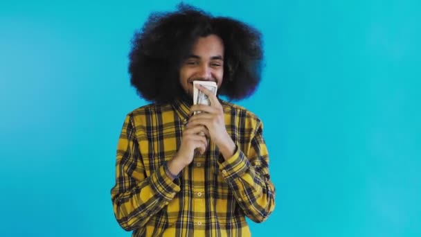Feliz joven afroamericano hombre sosteniendo el dinero en sus manos y mirando a la cámara en el fondo azul . — Vídeo de stock